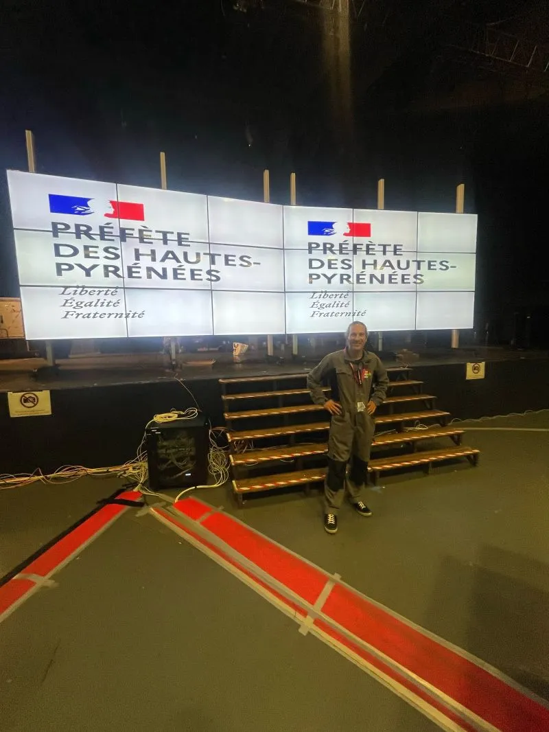Les deux murs de la salle affichant le logo de la préfète des Hautes-Pyrénées, un membre de l'équipe devant la scène